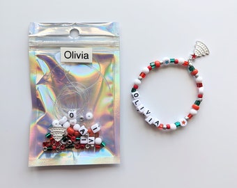 May include: A clear plastic bag with white beads, red and green beads, and a silver charm of a Christmas tree. The bag is labeled "Olivia". A white beaded bracelet with red and green beads and a silver Christmas tree charm is shown next to the bag. The bracelet has the name "Olivia" spelled out in white beads.