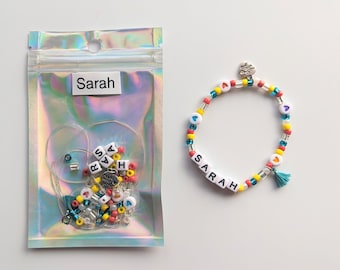 May include: A clear plastic bag with a white label that says "Sarah" and a variety of colorful beads, including letters, hearts, and stars. Next to the bag is a completed beaded bracelet with the name "SARAH" spelled out in white beads and a turquoise tassel.