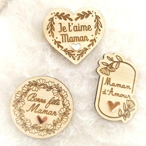 Op de afbeelding: Drie houten hartvormige ornamenten met bloemmotieven en de woorden "Je t'aime Maman", "Bonne f&ecirc;te Maman" en "Maman d'Amour". Elk ornament heeft een klein gouden hart in het midden.
