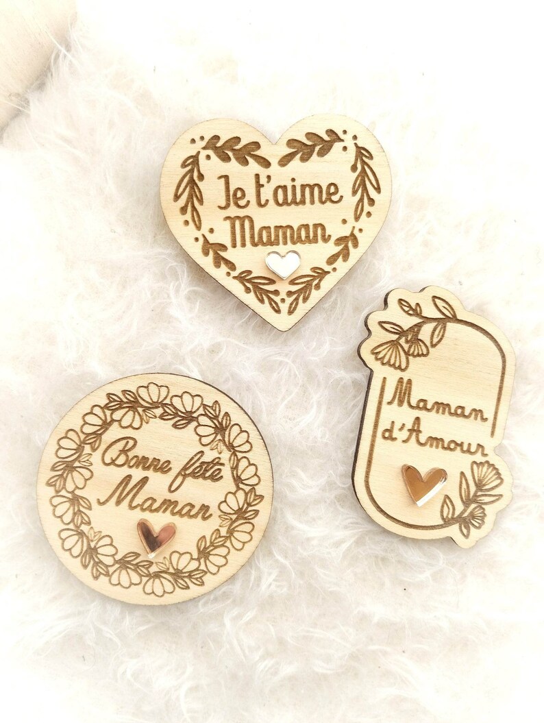 Op de afbeelding: Drie houten hartvormige ornamenten met bloemmotieven en de woorden "Je t'aime Maman", "Bonne f&ecirc;te Maman" en "Maman d'Amour". Elk ornament heeft een klein gouden hart in het midden.