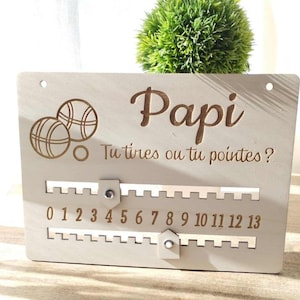 Peut inclure: Un tableau de score en bois pour un jeu de boules, avec le mot "Papi" et la phrase "Tu tires ou tu pointes?" Le tableau a un compteur coulissant avec les chiffres de 0 à 13.