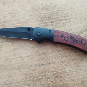 May include: A black folding knife with a wooden handle. The handle is engraved with the name "Franck".
