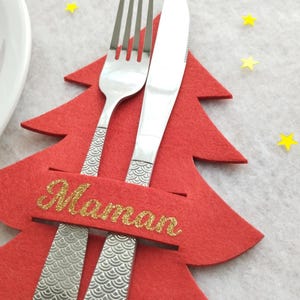 Puede incluir: Un portatarjetas con forma de árbol de Navidad de fieltro rojo con un tenedor y un cuchillo. El portatarjetas tiene la palabra "Maman" en purpurina dorada. Confeti amarillo en forma de estrella está esparcido.