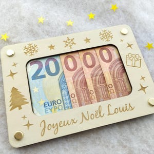 May include: A light wooden frame, decorated with engraved festive designs, holds Euro banknotes. The frame is inscribed with "Joyeux Noël Louis". Gold star-shaped confetti is scattered around the frame. The frame has a festive theme.