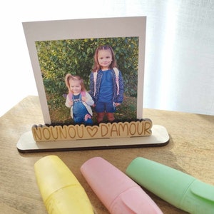 Peut inclure: Un porte-photo en bois avec le texte "NOUNOU ♥ D'AMOUR" et une photo de deux jeunes filles. Le porte-photo est posé sur une surface en bois avec trois surligneurs jaunes, roses et verts.