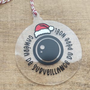May include: A clear, circular Christmas ornament featuring a black camera lens design and the French text "CAMÉRA DE SURVEILLANCE DU PÈRE NOËL". A Santa hat graphic sits above the lens. It hangs from a red and white string.