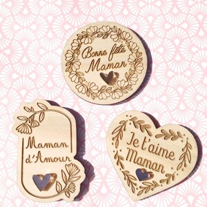 Op de afbeelding: Drie houten ornamenten met bloemmotieven en hartverwarmende boodschappen. De eerste ornament is rond en zegt "Bonne feste Maman". De tweede ornament is rechthoekig en zegt "Maman d'Amour". De derde ornament is hartvormig en zegt "Je t'aime Maman".