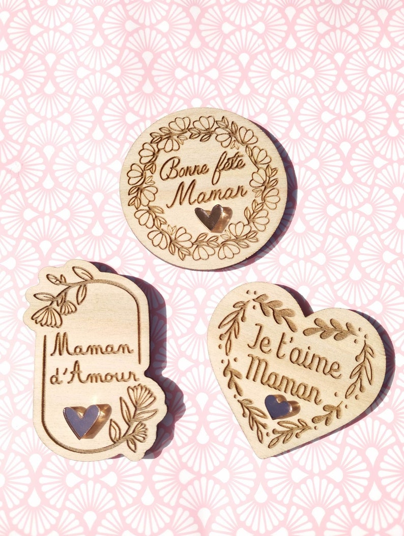 Op de afbeelding: Drie houten ornamenten met bloemmotieven en hartverwarmende boodschappen. De eerste ornament is rond en zegt "Bonne feste Maman". De tweede ornament is rechthoekig en zegt "Maman d'Amour". De derde ornament is hartvormig en zegt "Je t'aime Maman".