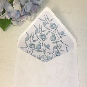 以下が含まれることがあります： 白い封筒で、内側のフラップに青い花柄が施されています。封筒は白い表面に平らに置かれています。