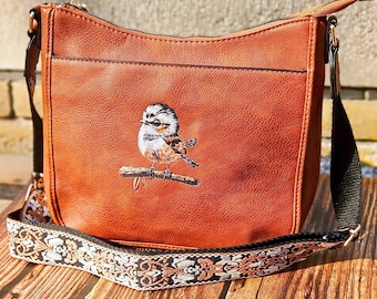 May include: A brown crossbody bag with an embroidered bird perched on a branch. The bag has a front pocket and a patterned strap with black and white floral designs. The bag is made of a leather-like material.