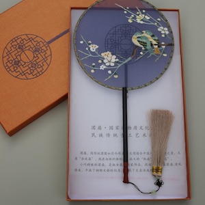 May include: A traditional Chinese hand fan with a blue silk fabric and embroidered flowers and birds. The fan is in a box with a description of the fan in Chinese characters.