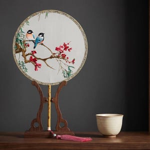 May include: A round, off-white embroidered fan featuring two birds perched on a flowering branch with red blossoms. The fan is displayed on a dark wooden stand with a tassel. A small, cream-colored ceramic cup sits nearby.