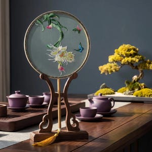 May include: A round, embroidered fan with a floral design and a wooden stand. The fan features a light green background with white flowers, pink accents, and a blue butterfly. The stand is dark brown with a gold tassel.
