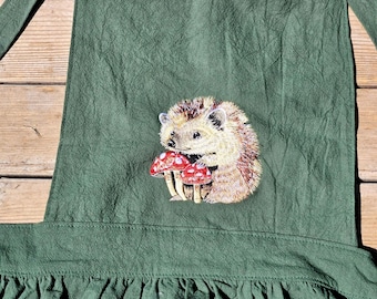 May include: A dark olive green apron featuring an embroidered design of a small brown hedgehog holding two red mushrooms with white spots. The apron has a gathered skirt section at the bottom and two long straps extending from the top corners.