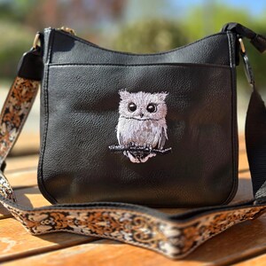 Könnte beinhalten: Schwarze Umhängetasche mit einem gestickten Eulen-Design. Die Tasche hat eine strukturierte Oberfläche und einen gemusterten Riemen in Gold-, Braun- und Cremetönen. Die Eule sitzt auf einem Ast, mit detaillierten Stickereien in Grau- und Weißtönen.