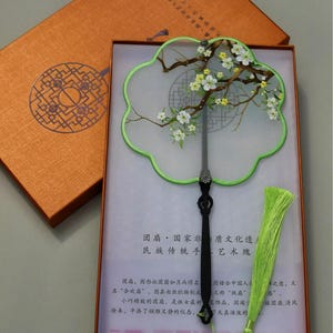 May include: An ornate, hand-painted fan with a green border and a black handle, presented in a decorative box. The fan features a floral design with white and yellow blossoms on brown branches. A green tassel is included.