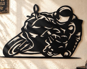 May include: A black metal wall art piece depicting a motorcycle rider leaning into a turn. The silhouette features a rider in a helmet and racing suit on a sportbike. The design is cut from a single sheet of metal with negative space details.