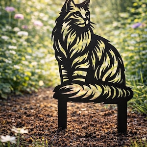 May include: Black metal cat silhouette garden stake. The cat is depicted in a sitting position with intricate cut-out details. The garden stake has two legs for easy placement in soil. The cat is set against a blurred background of greenery.