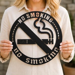 Op de afbeelding: Een rond zwart metalen bord met een uitgesneden ontwerp van een sigaret met opstijgende rook, doorgestreept door een diagonale lijn. De tekst NO SMOKING is in de boven- en onderkant van het ronde frame gesneden in een vet, gestileerd lettertype.