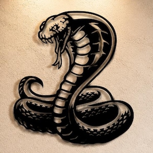 May include: Black metal cobra wall art. The snake is depicted with its hood flared and mouth open, ready to strike. The artwork is mounted on a light-colored wall, creating a striking contrast. This piece adds a bold, modern touch to any space.
