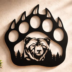 May include: Black metal bear paw wall art featuring a bear's head and a forest silhouette. The artwork is mounted on a neutral-colored wall above a wooden console table. The bear's head is detailed, and the paw has five distinct toe pads.