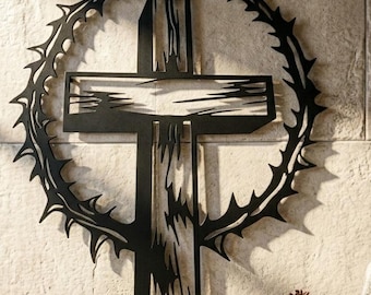 May include: A black metal wall art piece featuring a central cross design surrounded by a circular crown of thorns. The cross has a textured, distressed appearance with sharp, jagged edges, and the entire structure is mounted on a stone wall.