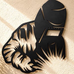 Op de afbeelding: Een zwart metalen wanddecoratie met een gestileerd silhouet van een lasser die een beschermende helm draagt. Het ontwerp bevat scherpe, hoekige uitsparingen die de vorm van de helm en de getextureerde, vloeiende lijnen van een baard definiëren.