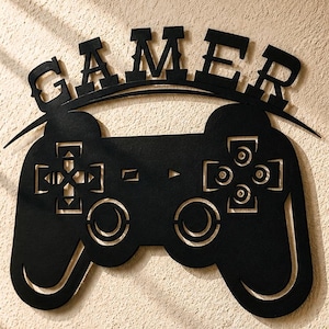 May include: A black metal wall sign featuring the word GAMER above a game controller silhouette. The setup includes black headphones on a stand, a small sign reading EAT SLEEP GAME REPEAT, a game controller, a figurine, and a black mug with heart icons.