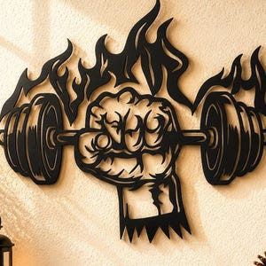 May include: A black metal wall art piece featuring a clenched fist gripping a barbell, surrounded by stylized flames. The design is cut from a single sheet of metal, showcasing bold lines and a silhouette of a hand lifting weights with fire.
