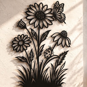 Op de afbeelding: Een zwart metalen wanddecoratie met een bloemmotief, bestaande uit twee grote madeliefachtige bloemen, een vlinder rechtsboven en een klein lieveheersbeestje op een blad. Het silhouet bevat stengels, bladeren en gras aan de onderzijde.