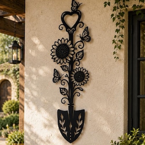 May include: A black metal wall art piece shaped like a garden shovel. The handle features a heart outline topped with a butterfly, while the shaft is decorated with two large sunflowers, swirling vines, leaves, and two additional butterflies.