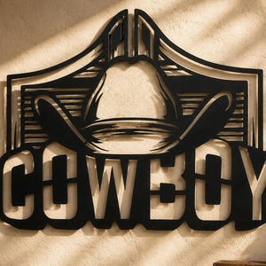 May include: A black metal wall art piece featuring a cutout design of a cowboy hat positioned above the word COWBOY in bold, capital letters. The design includes horizontal line accents behind the hat, creating a structured, decorative silhouette.