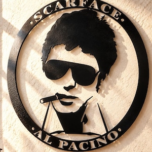 May include: A circular black metal wall art piece featuring a silhouette of a person wearing sunglasses and holding a cigar. The text SCARFACE is arched at the top and AL PACINO is arched at the bottom within the circular frame border.
