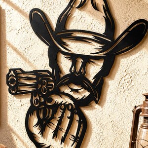 May include: A black metal wall art piece depicting a stylized cowboy wearing a wide-brimmed hat and a mustache, holding a revolver. The design features cut-out details and clean lines, showcasing a silhouette of a person in western attire.