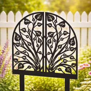 Puede incluir: Una estaca de jardín de metal negro con un diseño decorativo de hojas. La estaca tiene dos postes verticales y una parte superior semicircular con patrones de hojas y ramas recortadas. La estaca está colocada en un parterre con flores y una valla blanca al fondo.