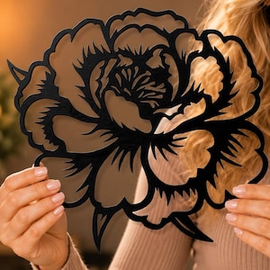 May include: Black laser-cut metal flower decoration. The floral design features intricate details and a layered appearance. The flower is held up in front of a neutral background. The flower's petals are detailed and the overall shape is round.