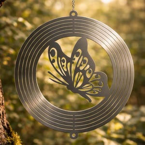 Pode incluir: Um móbile de vento de metal com uma borboleta pendurada por uma corrente. A borboleta é recortada do metal e é cercada por uma moldura circular com linhas concêntricas. O fundo é uma vista desfocada de uma floresta.