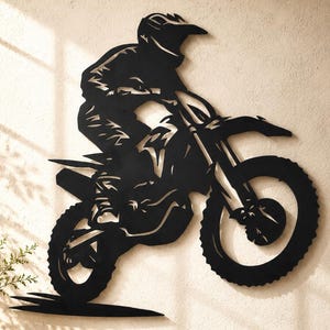 Może przedstawiać: Czarna metalowa dekoracja ścienna przedstawiająca sylwetkę motocyklisty crossowego na motocyklu w powietrzu. Projekt zawiera wycięte detale ukazujące kierowcę w kasku i stroju ochronnym na motocyklu w trakcie skoku.