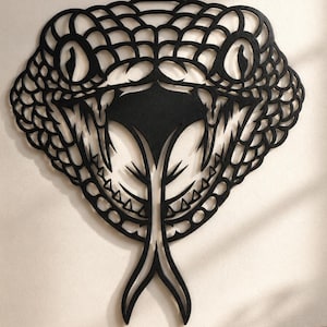 Puede incluir: Una escultura de pared negra con forma de cabeza de serpiente que presenta un patrón de escamas detallado, mandíbulas abiertas con colmillos visibles y una lengua bífida larga que se extiende hacia abajo. El diseño tiene recortes complejos.
