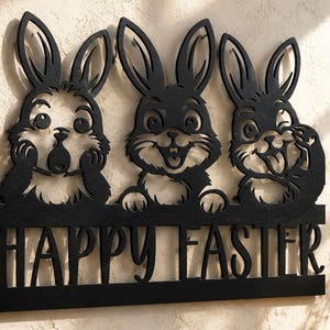Może przedstawiać: Czarny metalowy znak ścienny przedstawiający trzy króliki z kreskówek o różnych wyrazach twarzy, umieszczone nad napisem HAPPY EASTER. Króliki mają długie uszy i szczegółowe tekstury futra, a całość jest płaskim wycięciem.