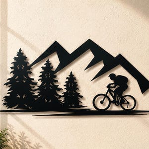 Könnte beinhalten: Ein Wandkunstobjekt aus schwarzem Metall, das die Silhouette eines Gebirges, drei Tannen und eine Person auf einem Mountainbike zeigt. Das Design ist flach aus einem Stück Material geschnitten und weist scharfe Kanten sowie ein dunkles Finish auf.