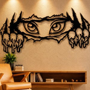 Op de afbeelding: Een zwart metalen wandkunstwerk dat de ogen van een wezen toont dat door een gescheurd oppervlak kijkt, met klauwmarkeringen die naar buiten uitsteken. Het kunstwerk is gemonteerd op een beige muur, boven een houten kast en een leren stoel.