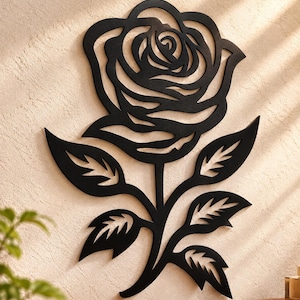 May include: A black metal wall art piece shaped like a rose with a detailed bloom and a stem featuring four pointed leaves. The design is a flat, cutout silhouette that displays the intricate curves of the petals and the serrated edges of the foliage.