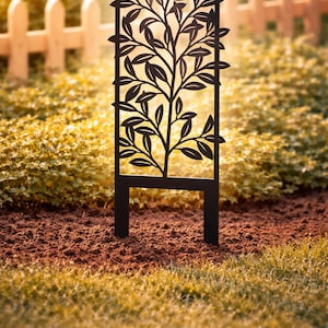 Puede incluir: Un enrejado de jardín de metal negro con un diseño de árbol. El enrejado presenta un marco rectangular con una silueta recortada de un árbol con hojas. Tiene dos patas para colocarlo en el suelo, sobre un fondo de jardín.
