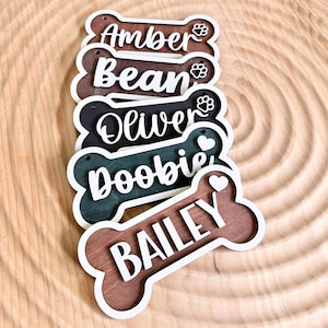 May include: A stack of wooden dog bone-shaped name tags in various colors, including brown, black, and teal. Each tag features a different dog's name in white script, with paw prints or hearts. The tags are layered with a white border.