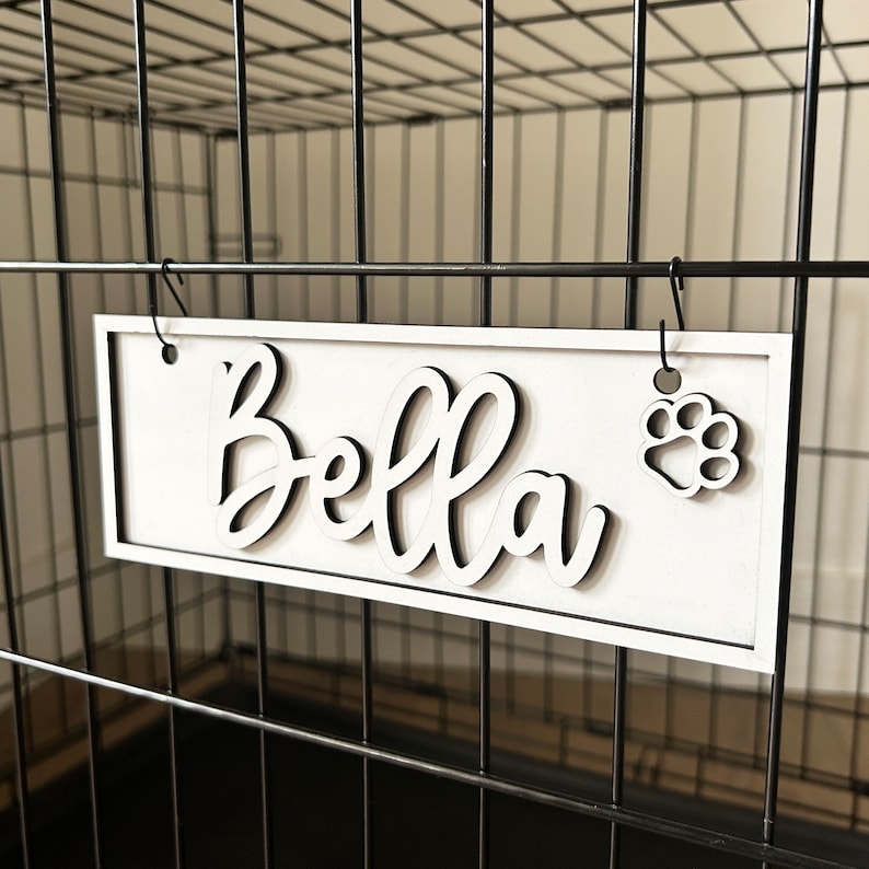 K&ouml;nnte beinhalten: Ein wei&szlig;es Holzschild mit dem Namen "Bella" in schwarzer Kursivschrift und einem schwarzen Pfotenabdruck. Das Schild h&auml;ngt an einem schwarzen Metallhundek&auml;fig.