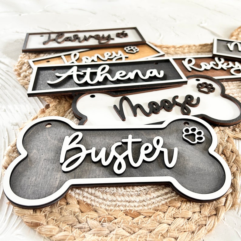 K&ouml;nnte beinhalten: Eine Sammlung personalisierter, holzspezifischer Hunde-Knochen-Etiketten mit Namen wie "Buster", "Moose", "Athena", "Honey" und "Rocky". Die Etiketten sind in einem dunklen Grau mit wei&szlig;en Buchstaben und einem Pfotenabdruck-Design bemalt.