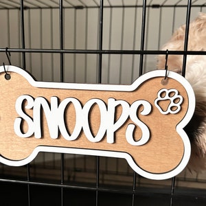 Könnte beinhalten: Ein hölzernes, knochenförmiges Schild für einen Hundekäfig mit dem Namen "Snoops" in weißen Kursivbuchstaben und einem weißen Pfotenabdruck. Das Schild hängt an der Oberseite eines schwarzen Metallhundekäfigs.