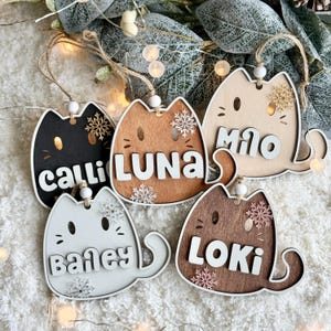 Könnte beinhalten: Fünf hölzerne Weihnachtsornamente in Katzenform. Jedes Ornament hat eine andere Farbe und einen anderen Namen: Calli, Luna, Milo, Bailey und Loki. Die Ornamente weisen Schneeflockenmuster auf und sind mit Bindfäden zum Aufhängen versehen. Eine festliche Weihnachtsdekoration.