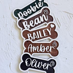 May include: Wooden dog name tags in a bone shape, with white trim. The tags display names in white script: Doobie, Bean, Bailey, Amber, and Oliver. Some tags have paw prints or a heart symbol.